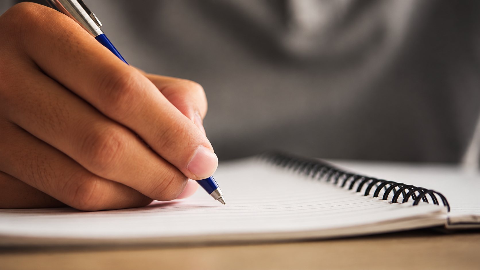 Gros plan d'une main tenant un stylo bleu et écrivant sur un carnet à spirale posé sur une table.